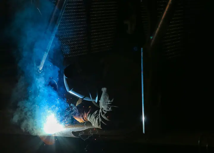 Welder in dark workshop with blue-tinged arc smoke, face fully shielded
