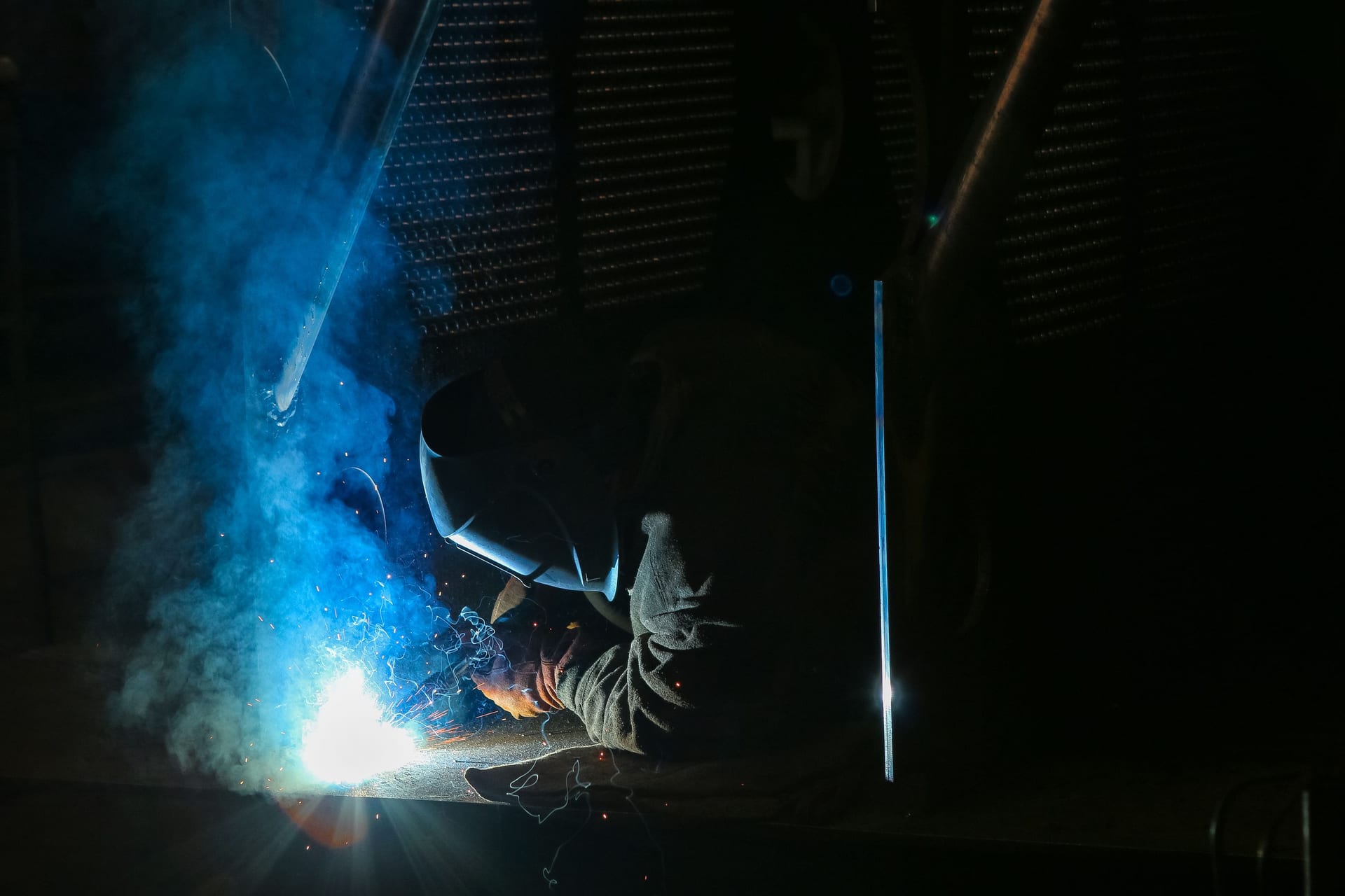 Welder in dark workshop with blue-tinged arc smoke, face fully shielded